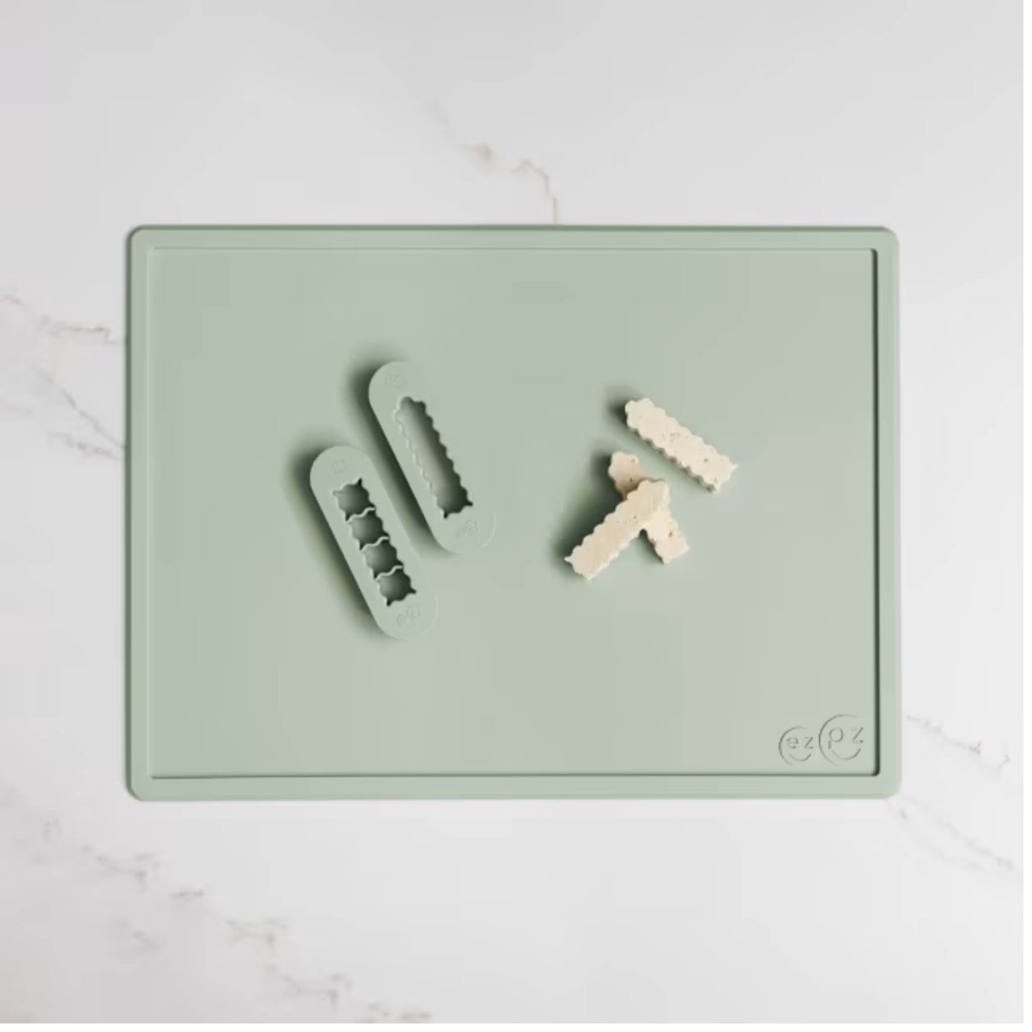 Sage green silicone food cutters beside small cubes of soft food prepared for baby finger foods on a matching cutting board.
