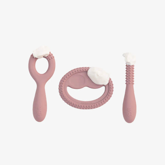 Three blush pink silicone oral development tools shown with food placed on surfaces demonstrating textured bumps and shapes designed to support early chewing skills.