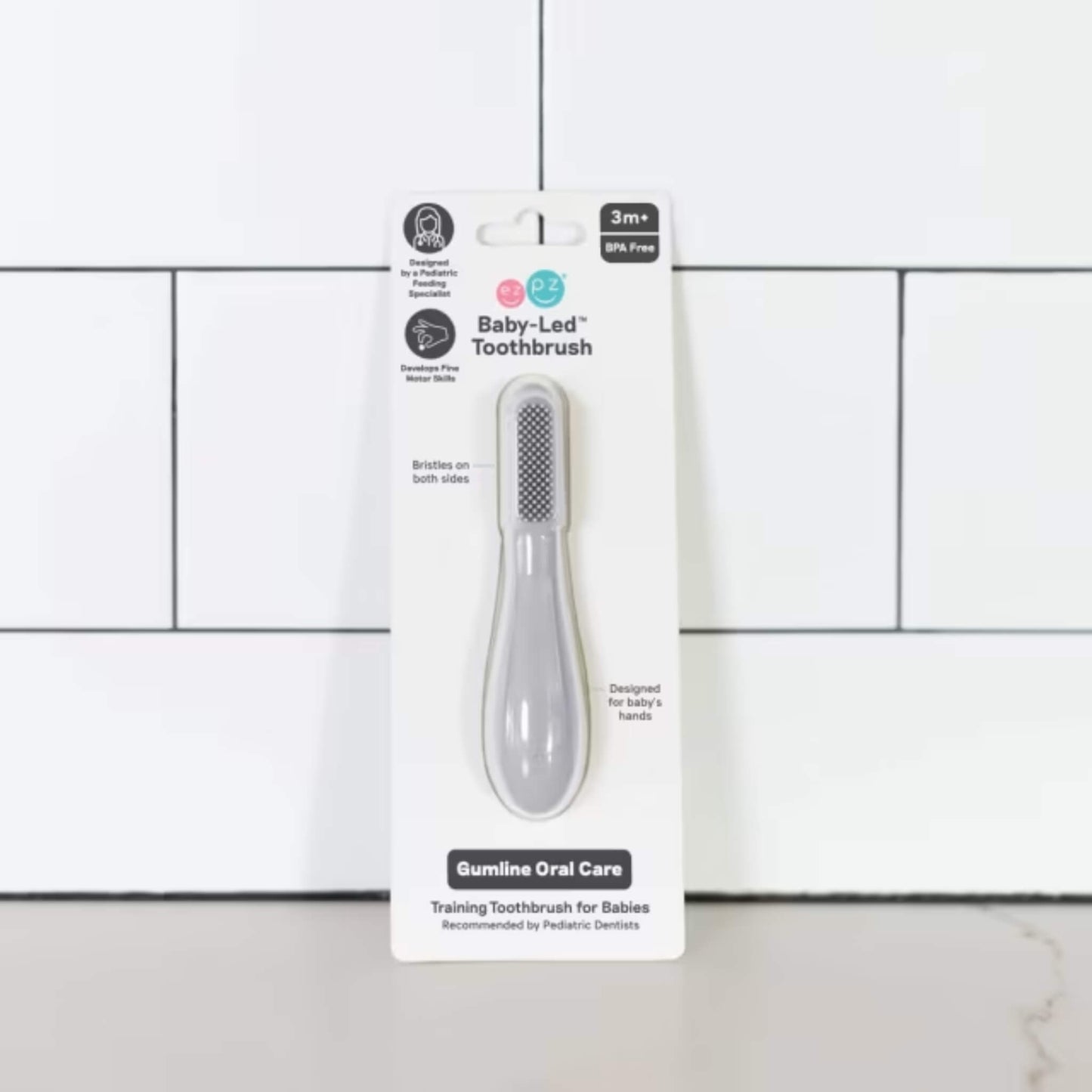 Grey silicone baby toothbrush displayed in retail packaging labelled baby-led toothbrush for babies 3+ months with dual-sided bristles and gumline oral care design.