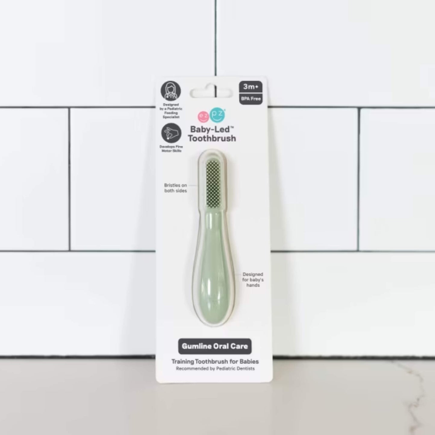 Sage green silicone baby toothbrush displayed in retail packaging labelled baby-led toothbrush for babies 3+ months with dual-sided bristles and gumline oral care design.