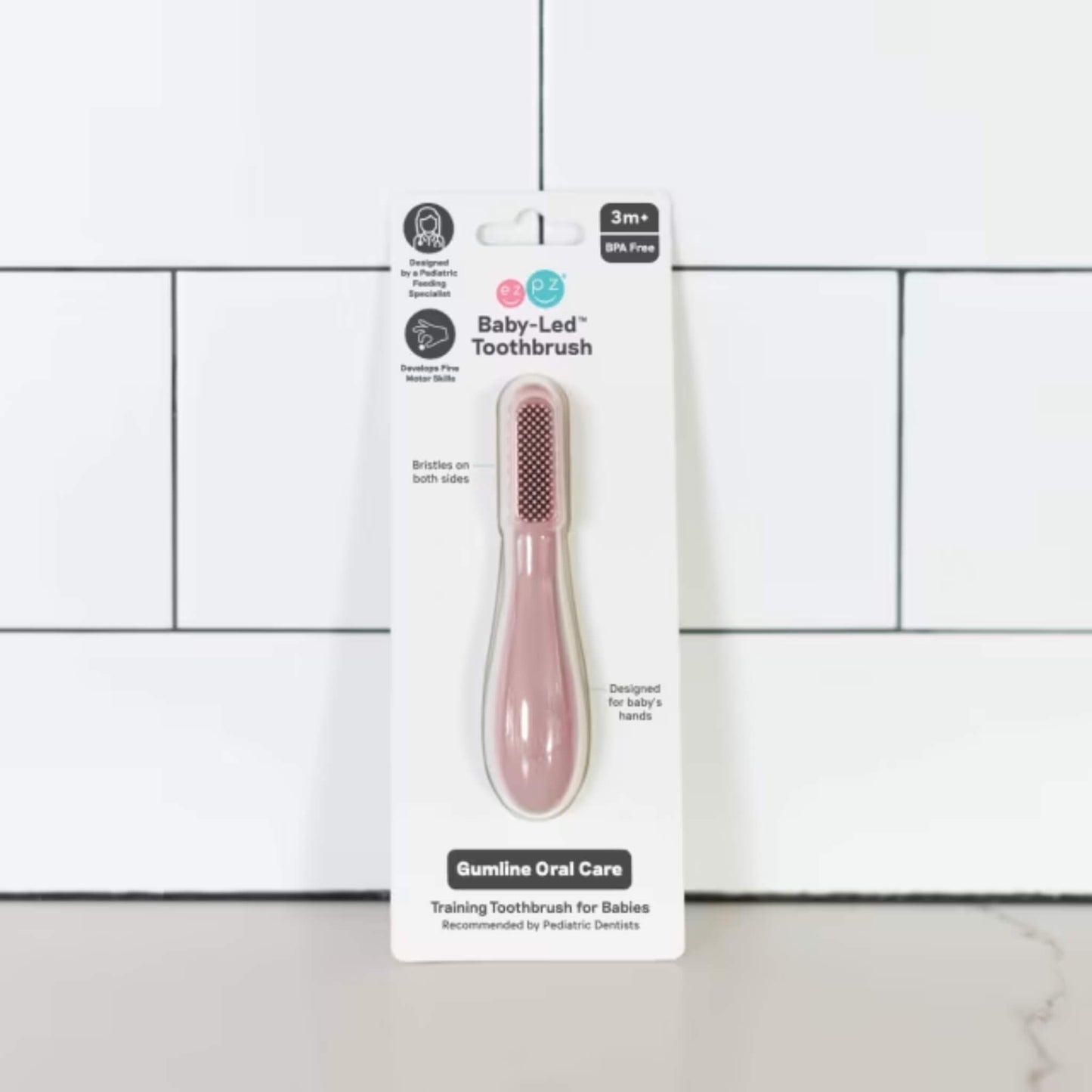 Silicone baby toothbrush in blush pink displayed in retail packaging labelled baby-led toothbrush for babies 3+ months with dual-sided bristles and gumline oral care design.