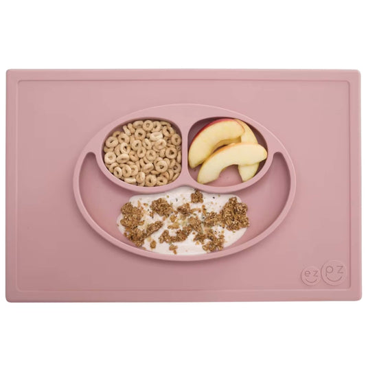 A blush pink silicone placemat and plate set with three compartments neatly filled with apple slices, cereal and yoghurt topped with granola.