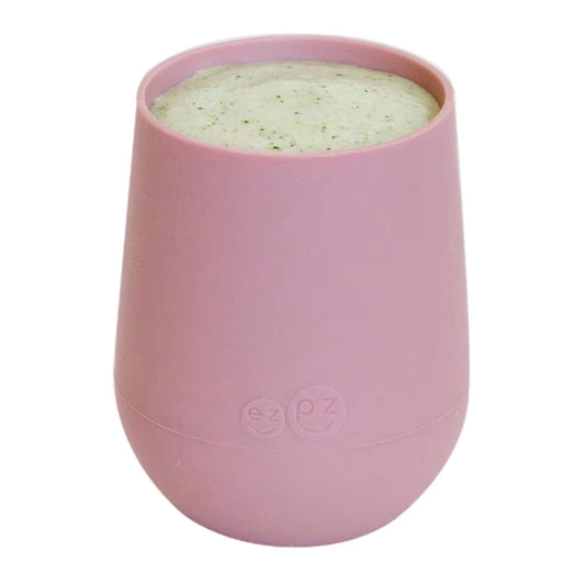 A blush pink silicone training cup shown filled with milk, designed for toddlers learning to drink from an open cup.