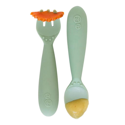 Sage green toddler training fork holding a slice of carrot beside a matching spoon filled with puree.