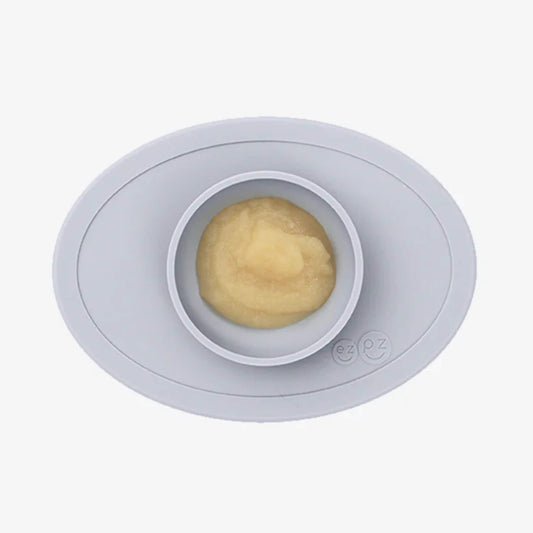 A grey silicone placemat with a small round bowl filled with smooth yellow puree, shown from above.