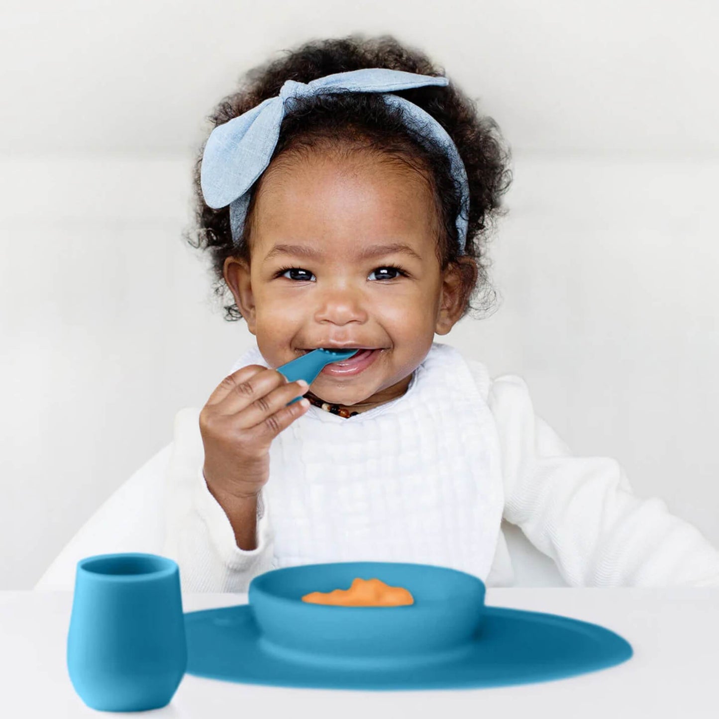Baby wearing a white bib and blue headband eating puree with a blue feeding set that includes a suction bowl, training spoon and open cup.