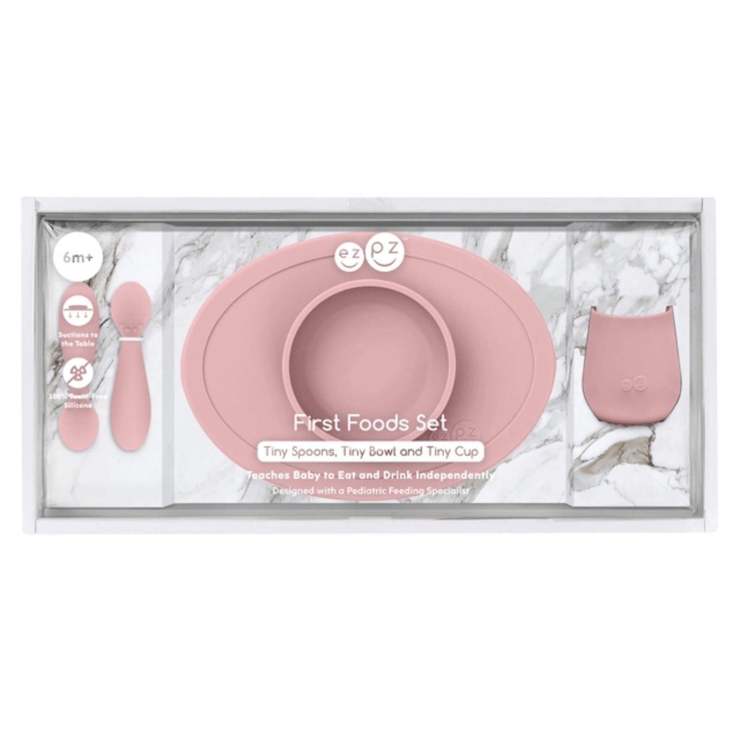 A baby first foods set in a clear plastic tray with a white marble-patterned insert. The set includes a dusty pink silicone bowl with a matching rim, two small pink spoons, and a small pink cup.