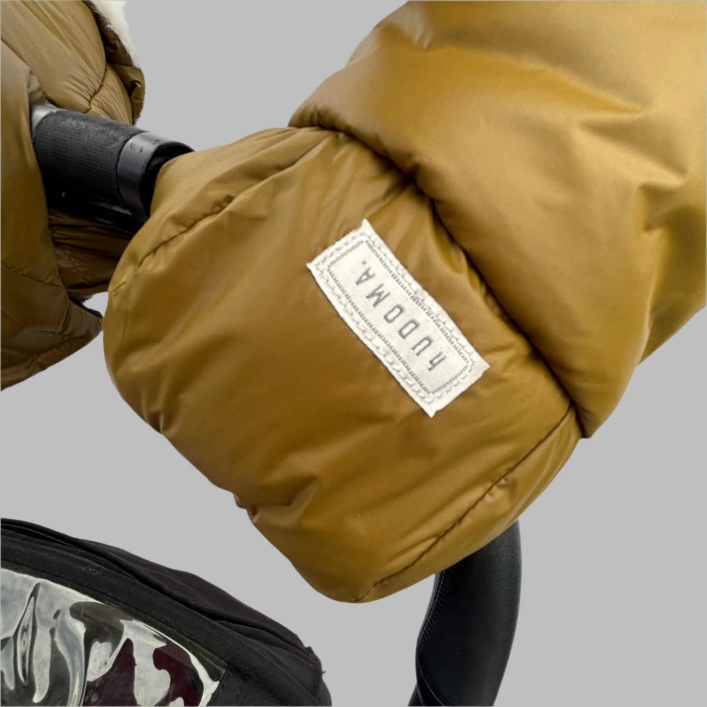 Close-up of green pram glove with stitched hUDOMA brand label attached to a stroller handle near a transparent rain cover.