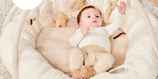 Baby lying on a soft padded Nattou playmat reaching up during tummy time, surrounded by plush neutral fabrics in a calm nursery setting.