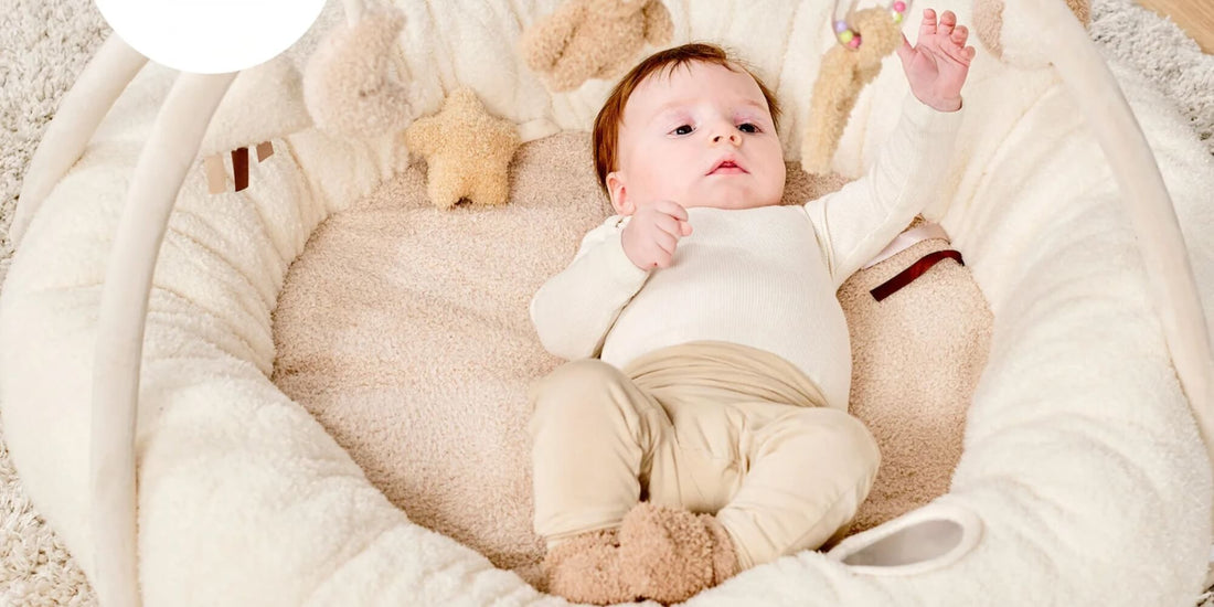 Baby lying on a soft padded Nattou playmat reaching up during tummy time, surrounded by plush neutral fabrics in a calm nursery setting.
