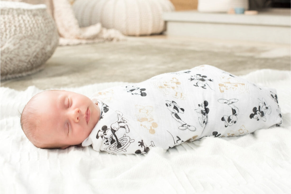 Newborn baby sleeping swaddled in a white muslin wrap with Mickey Mouse print, lying on a soft white blanket in a calm neutral-toned nursery.