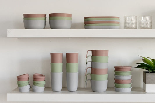 Pastel pink, sage green and pale grey silicone bowls, plates, cups and snack pots stacked neatly on white kitchen shelves beside two glass tumblers and a small plant.