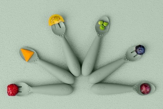 Six sage green ezpz Tiny Utensils arranged in a circle, each holding a small piece of fruit, vegetable or cheese.