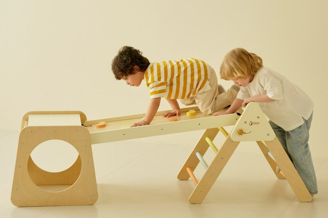 Two young children climbing across a wooden indoor climbing frame with a ramp and ladder, using their hands and knees to balance and move along the structure.