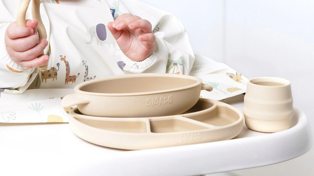 Baby reaching for beige silicone weaning dishes on a highchair tray.