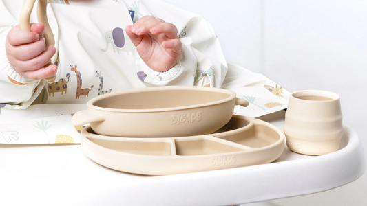 Baby reaching for beige silicone weaning dishes on a highchair tray.