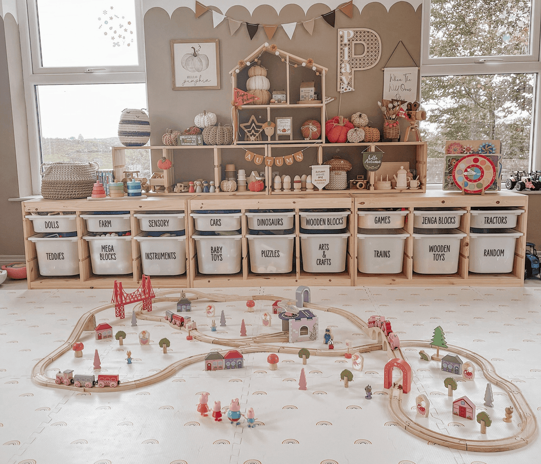 Wooden train track layout with small houses and figures set up on a playroom floor, with shelves of neatly labelled storage boxes and toys in the background.