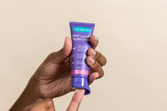 Lansinoh HPA Lanolin Nipple Cream tube held in a woman's hand, with a small amount of cream dispensed onto a fingertip against a neutral beige background.