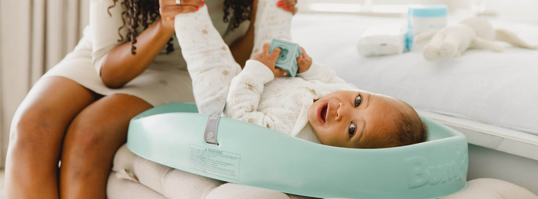 A smiling baby lies on a Hemlock Green Bumbo changing pad while a mother gently holds their legs, with the Bumbo logo subtly embossed on the side.