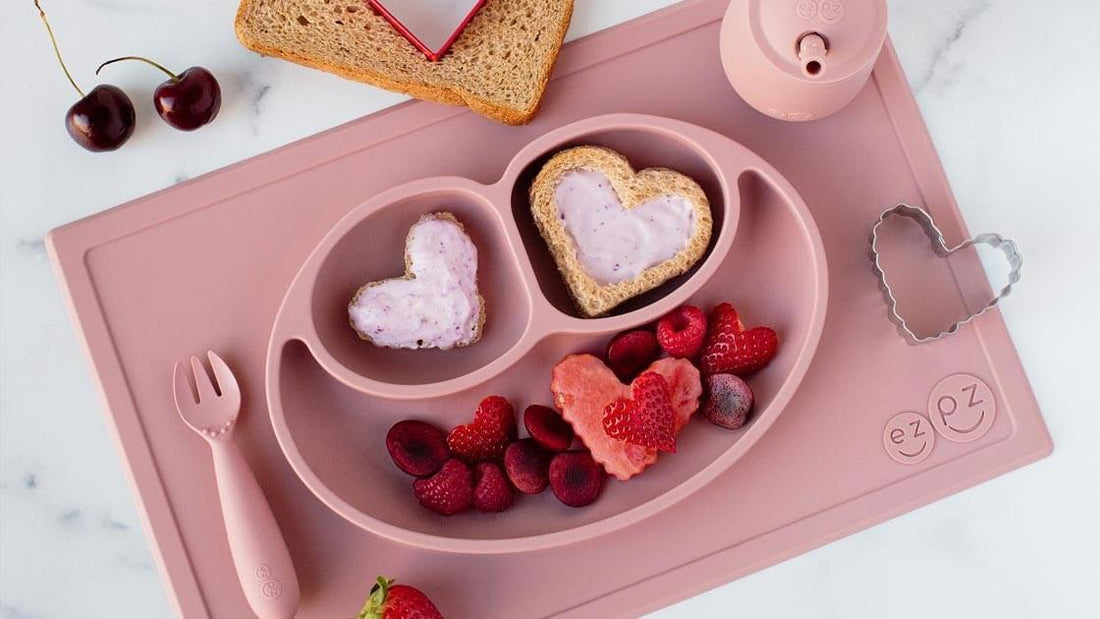 Pink silicone placemat and divided plate holding raspberries and heart-shaped waffles, with a matching spoon, open bowl and scattered fruit on a white surface.