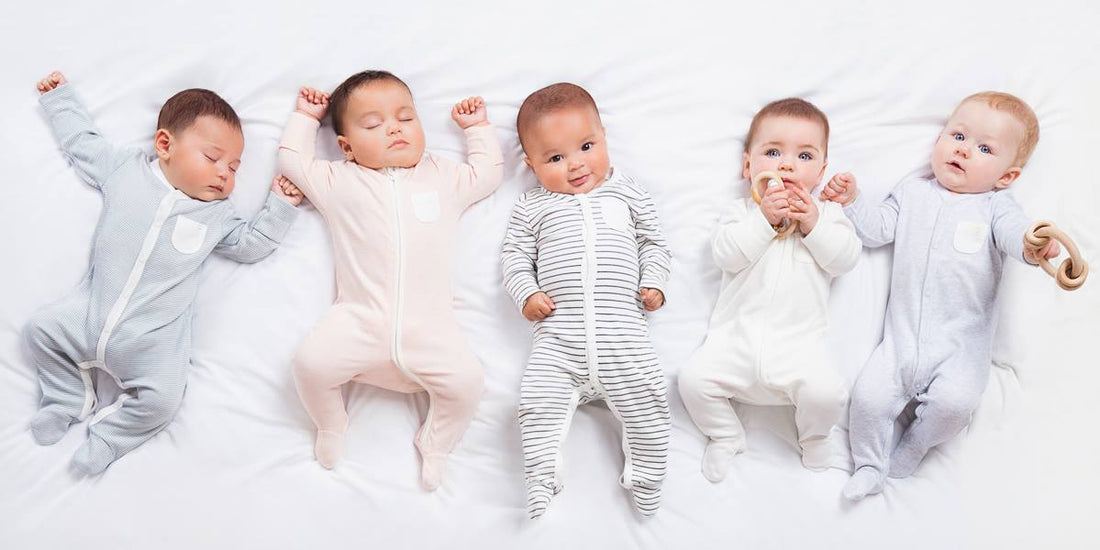 Five babies lying side by side on a white blanket, dressed in pale sleepsuits.