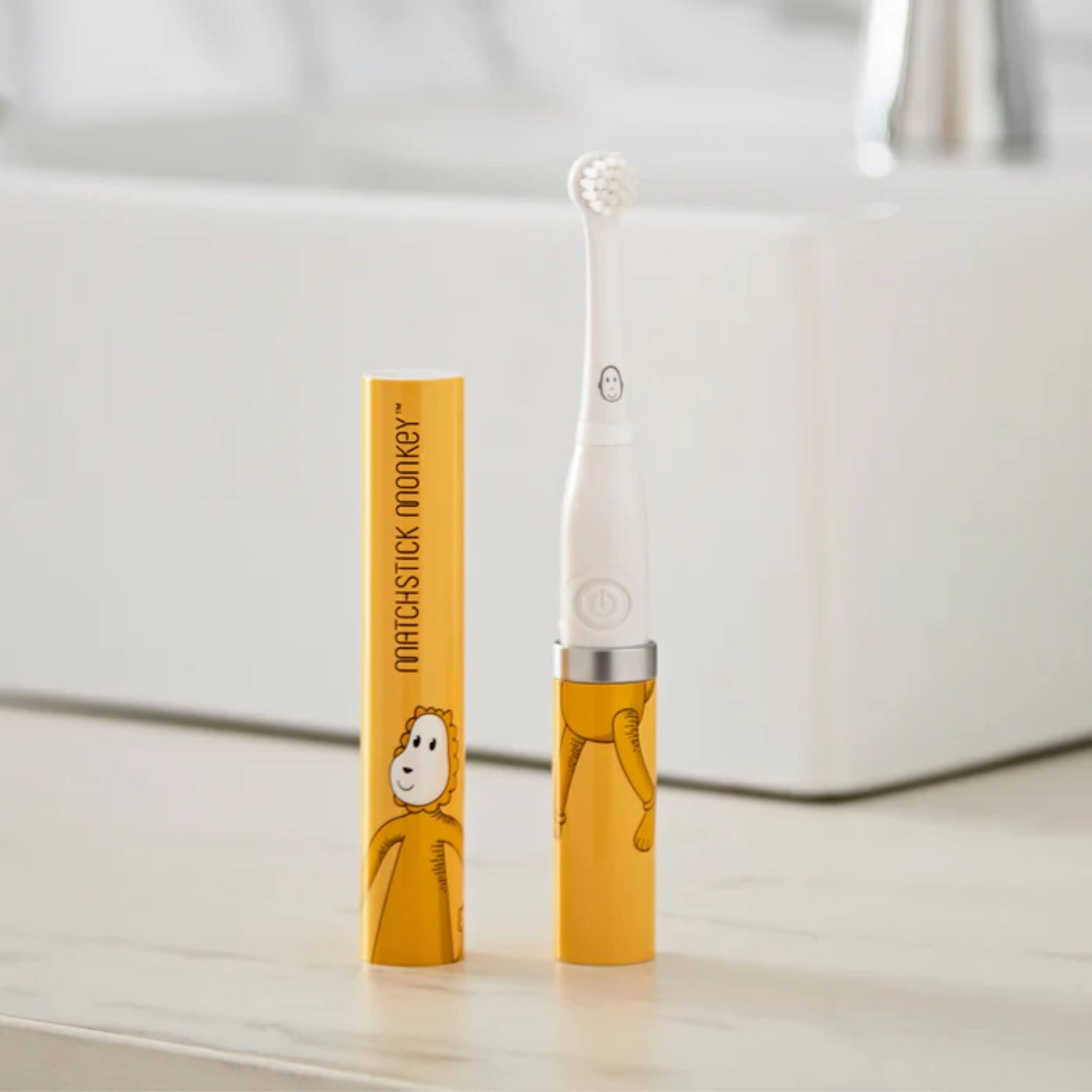 Electric sonic baby toothbrush with small soft bristle head standing upright beside a yellow illustrated protective travel case on a bathroom counter.