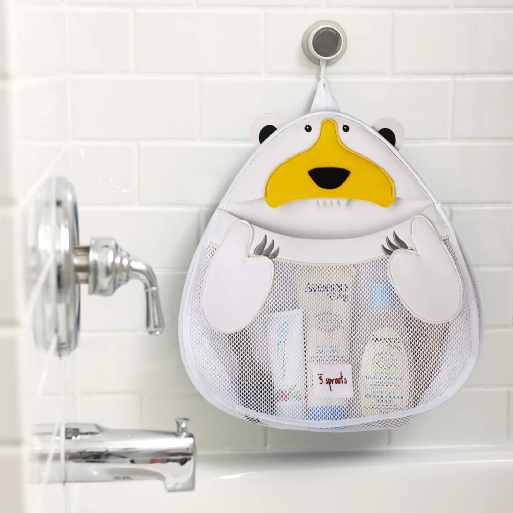 Polar bear bath storage organiser in use, attached to tiled wall above bathtub and filled with baby bath products in the mesh pouch.