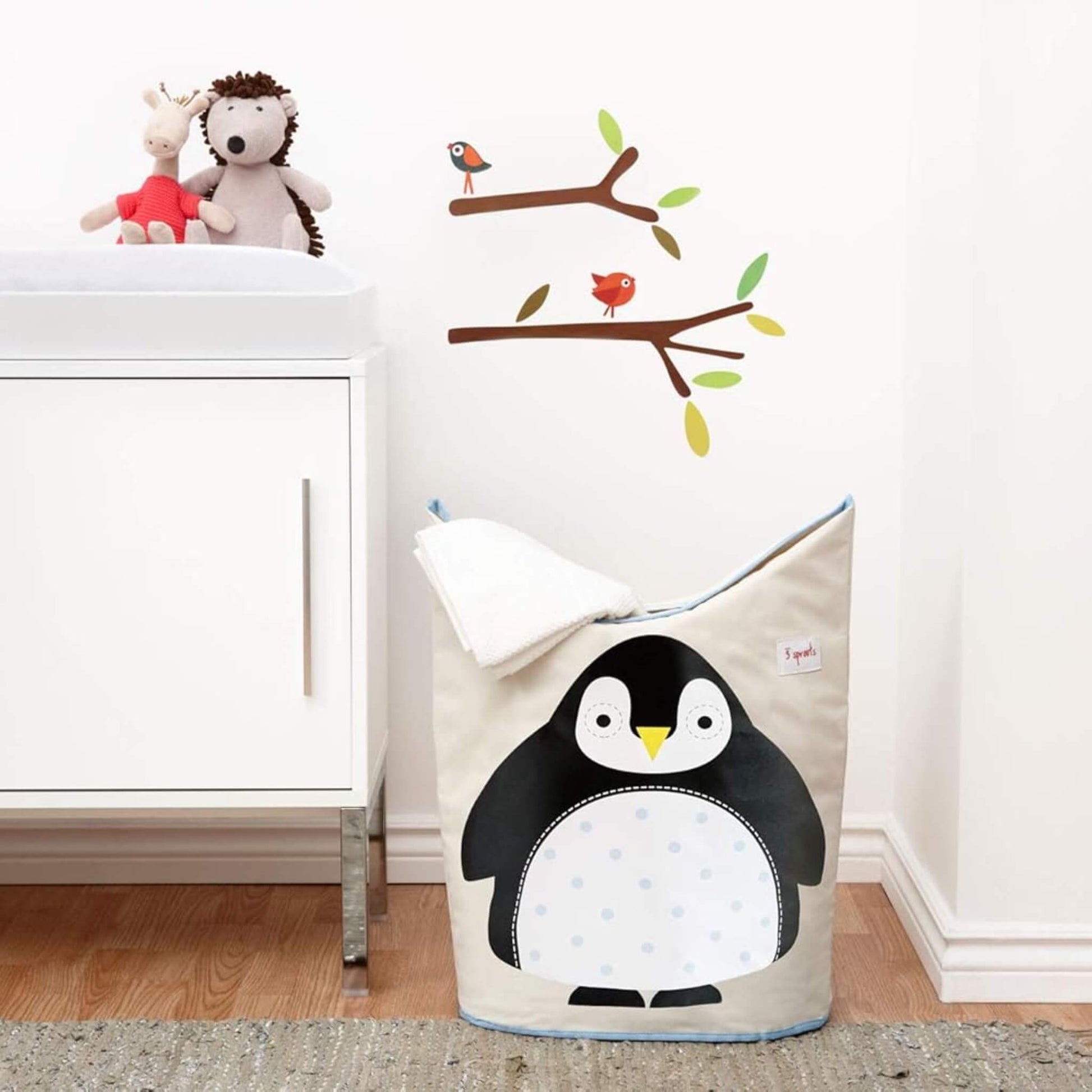 Penguin laundry hamper placed in a nursery setting beside a changing unit, with towel draped over top for real-life context.