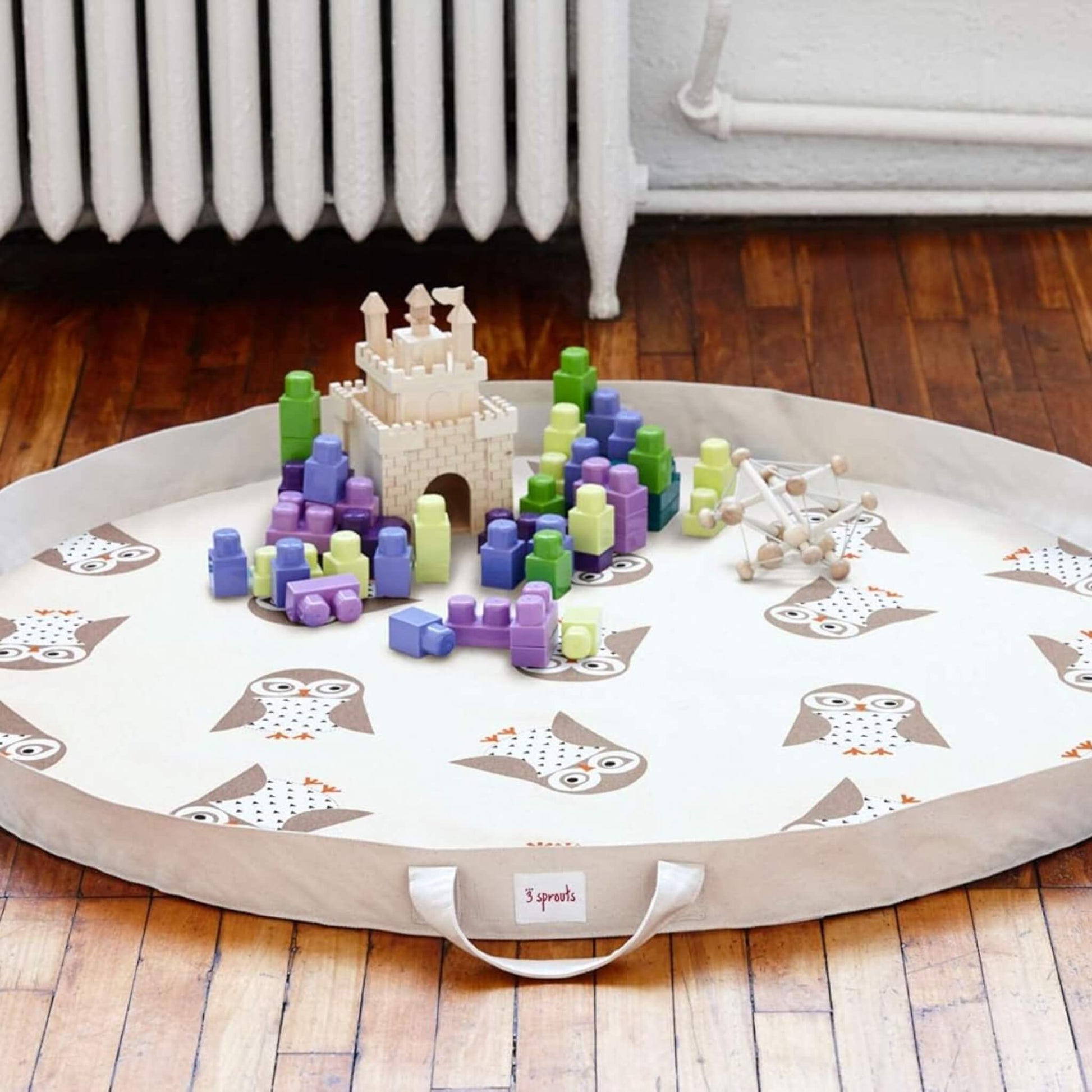 Owl play mat laid flat on wooden floor, scattered with colourful toy blocks and a toy castle. Handle and 3 Sprouts logo facing forward.