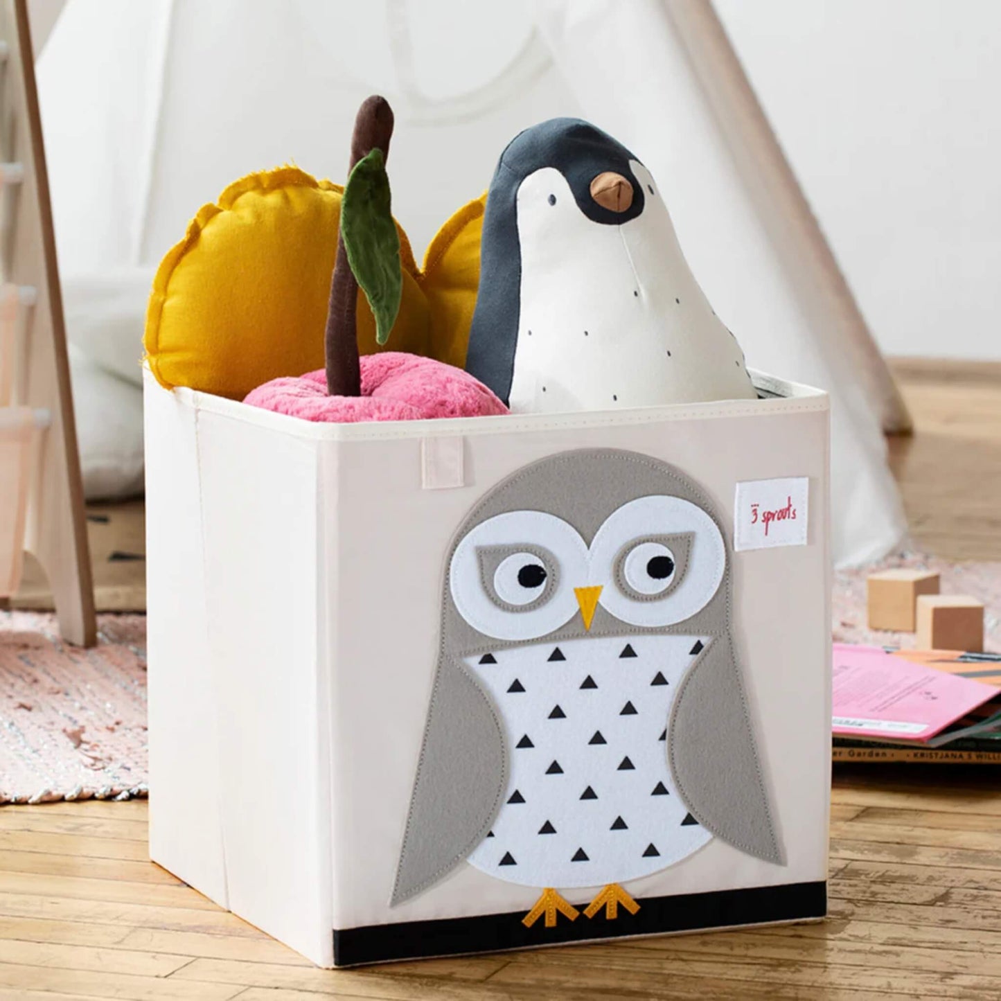 Close-up of the owl box filled with toys including a penguin soft toy and a yellow heart cushion, set on a wooden floor.