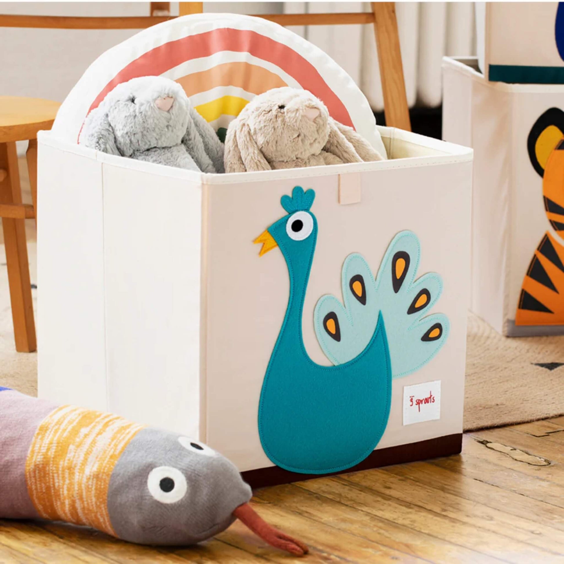 Peacock-themed storage box placed on a wooden floor, filled with soft toys and a rainbow cushion, next to other animal storage cubes.