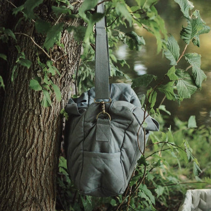 A quilted grey canvas baby changing bag hanging by its shoulder strap from a tree branch, showing soft structure and side pocket.