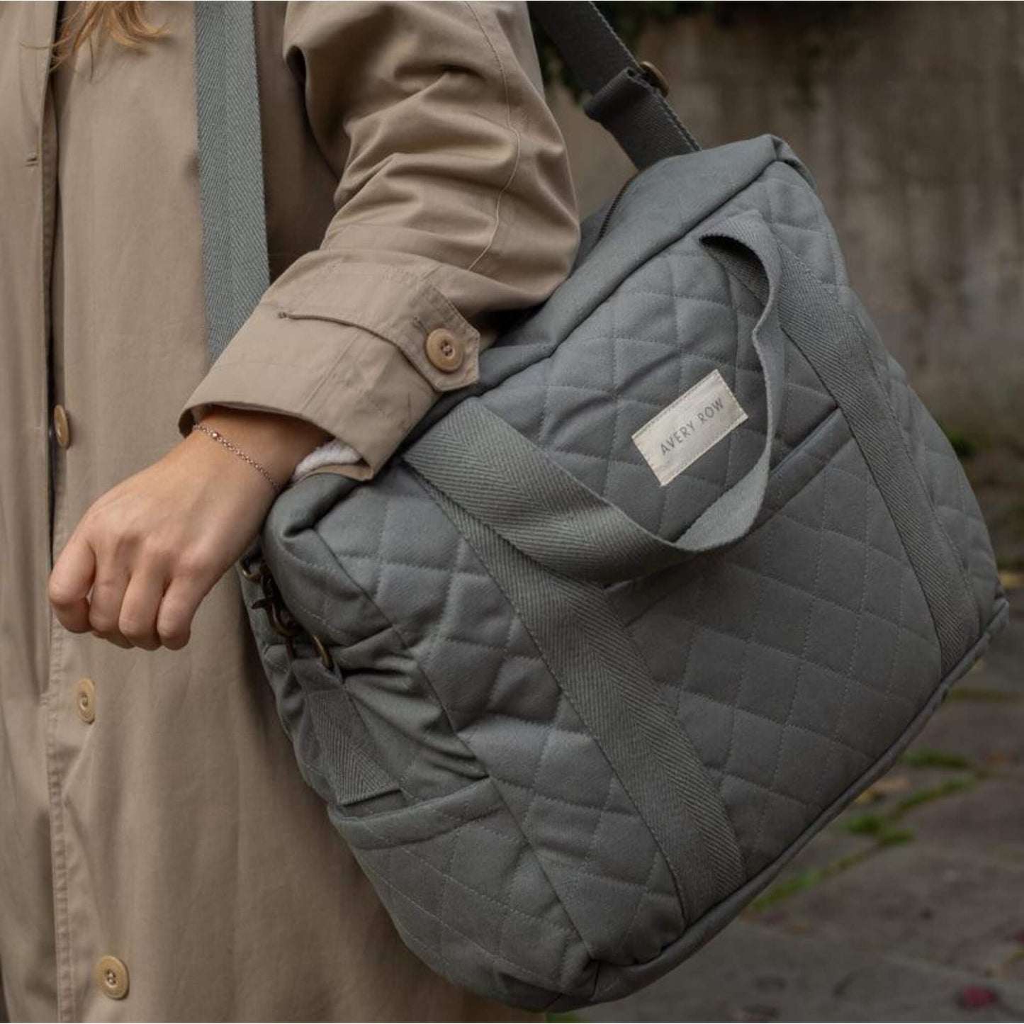 A quilted grey canvas baby changing bag carried on the shoulder, showing padded structure, stitched detailing and sturdy woven handles.