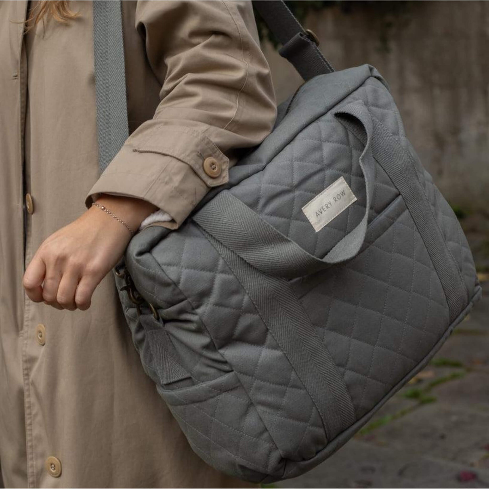 A quilted grey canvas baby changing bag carried on the shoulder, showing padded structure, stitched detailing and sturdy woven handles.