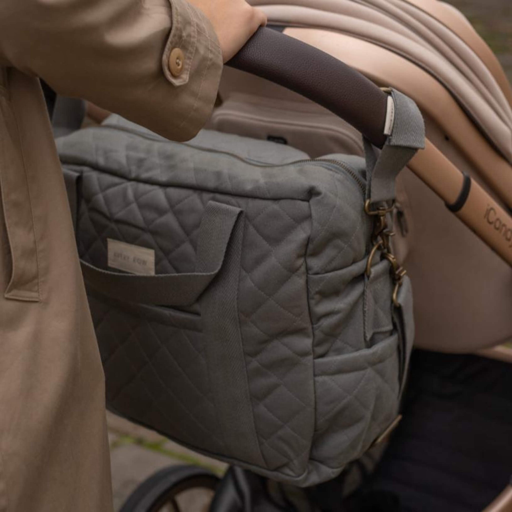A side view of a quilted grey canvas baby changing bag showing an external pocket and structured shape.
