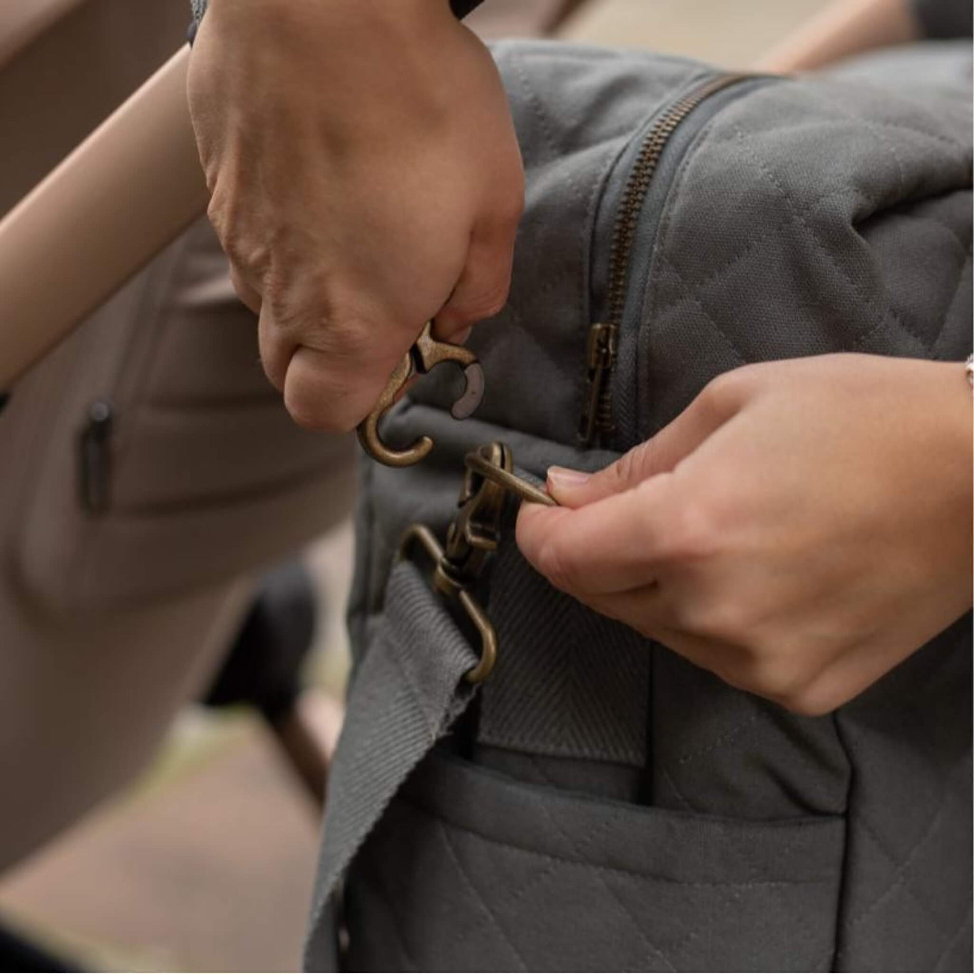 Hands attaching a detachable fabric strap to metal hardware on a quilted grey canvas baby changing bag.