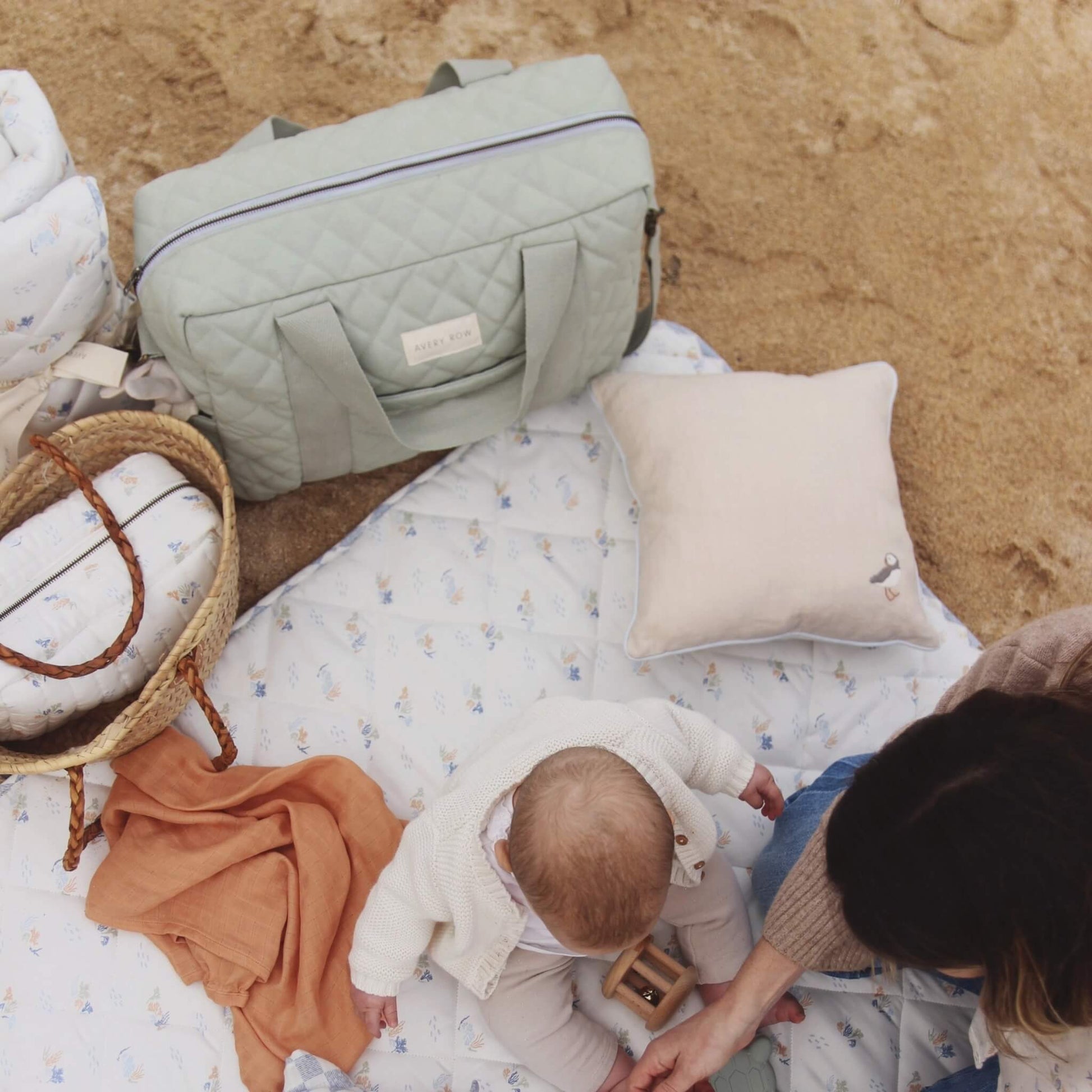 Overhead view of a baby sitting on a padded play mat at a sandy beach with a quilted changing bag, cushion, basket and blankets arranged nearby.