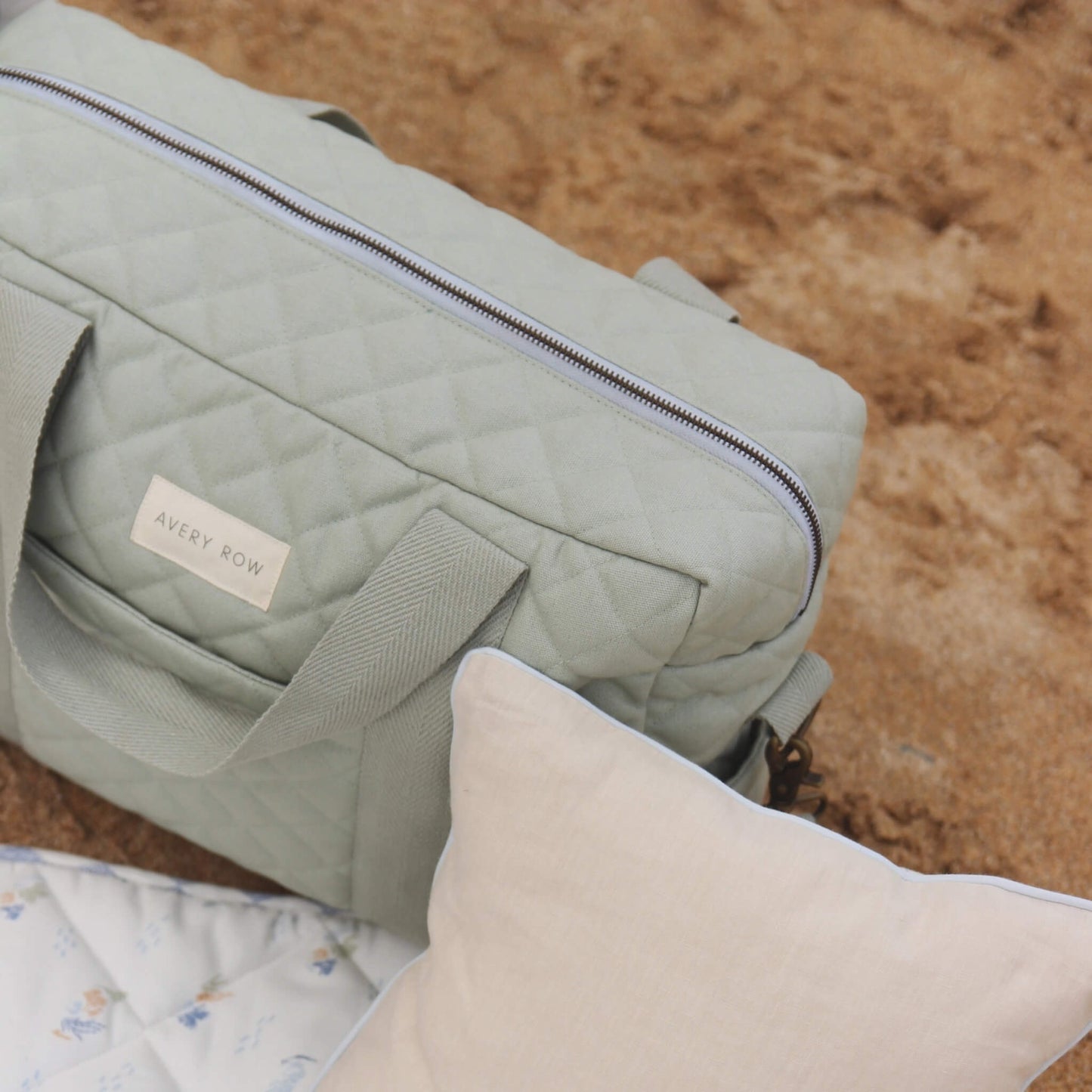 Sage green quilted changing bag resting on a picnic mat beside a neutral cushion, showing zip opening, handles and textured cotton fabric against sand.
