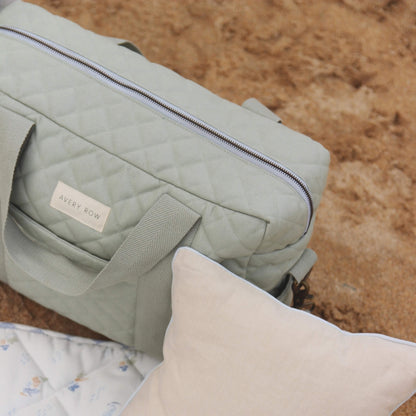 Sage green quilted changing bag resting on a picnic mat beside a neutral cushion, showing zip opening, handles and textured cotton fabric against sand.