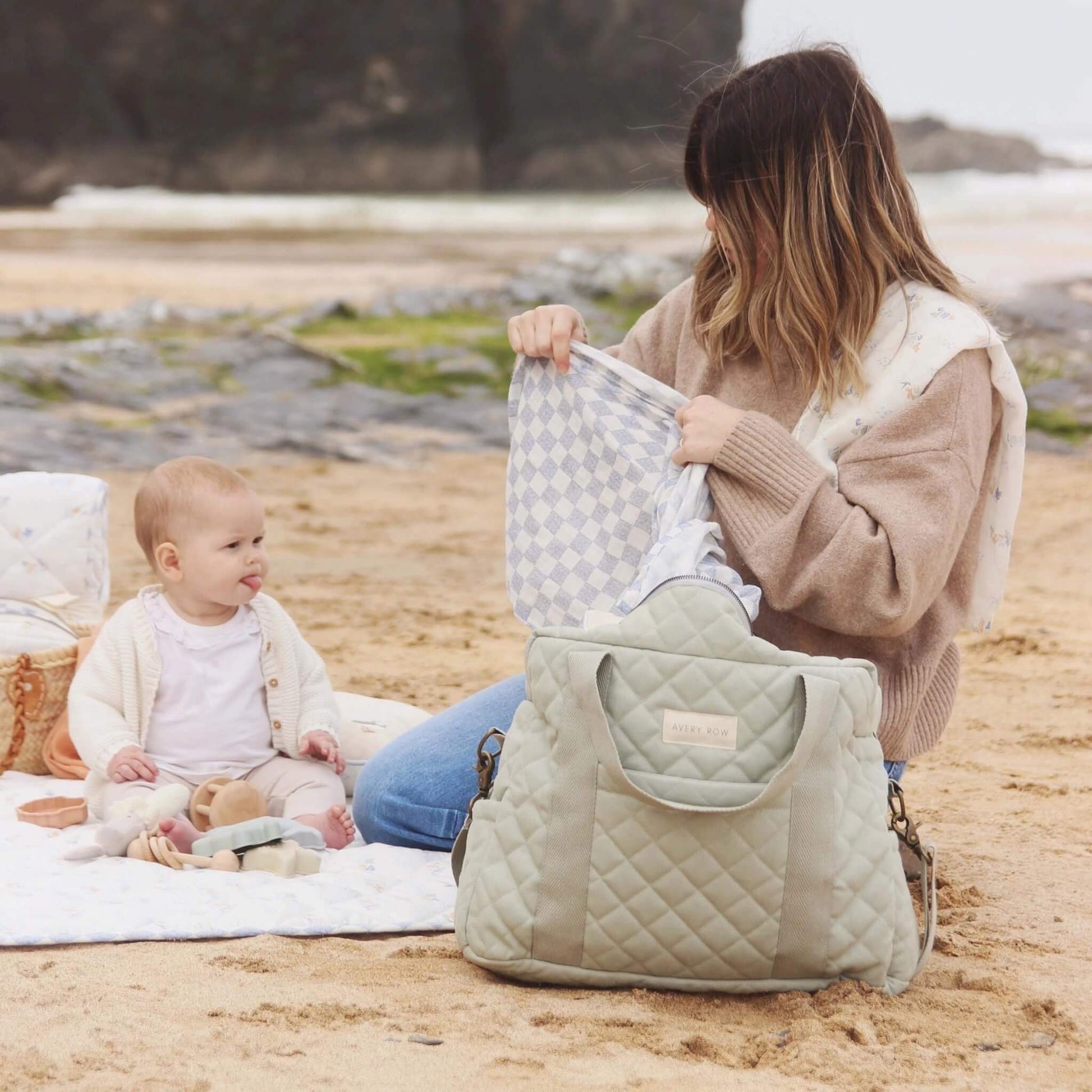 Woman sitting on a beach blanket placing a folded quilted mat into an open changing bag while a baby sits nearby, showing roomy interior and wide zip access.