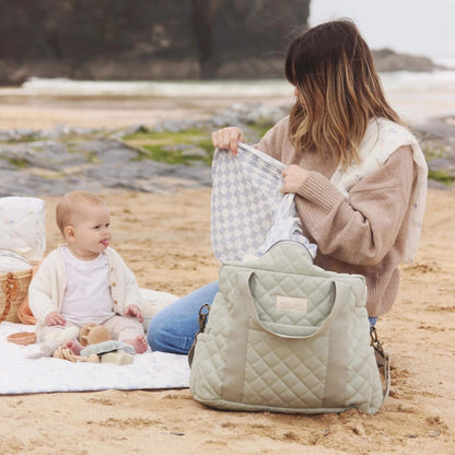 Woman sitting on a beach blanket placing a folded quilted mat into an open changing bag while a baby sits nearby, showing roomy interior and wide zip access.