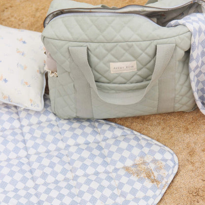 Front view of an open quilted changing bag on a picnic mat with lining visible inside, padded handles and structured shape shown in natural outdoor light.