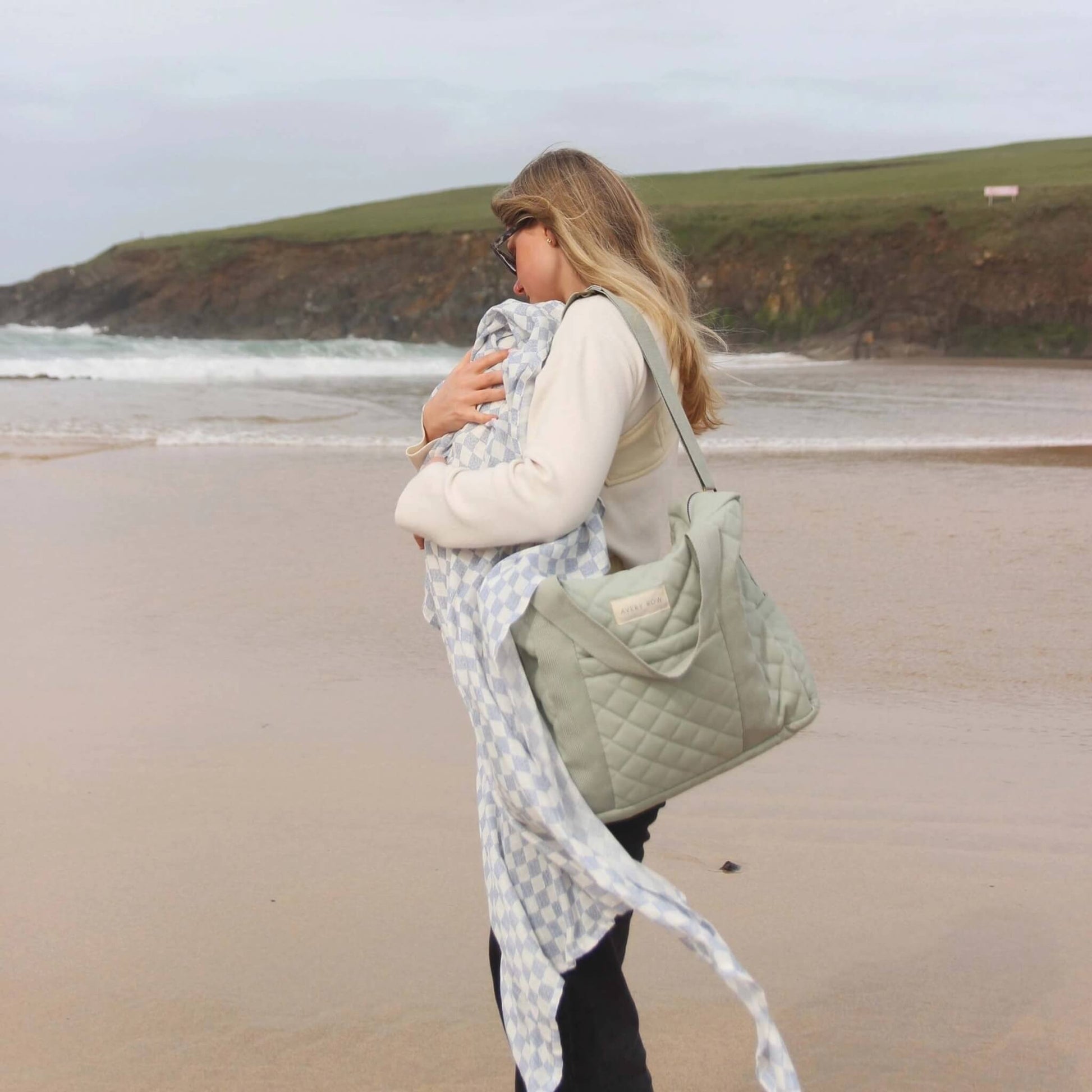 Woman walking along a sandy beach carrying a quilted sage green changing bag over her shoulder while holding a baby wrapped in a soft patterned blanket.