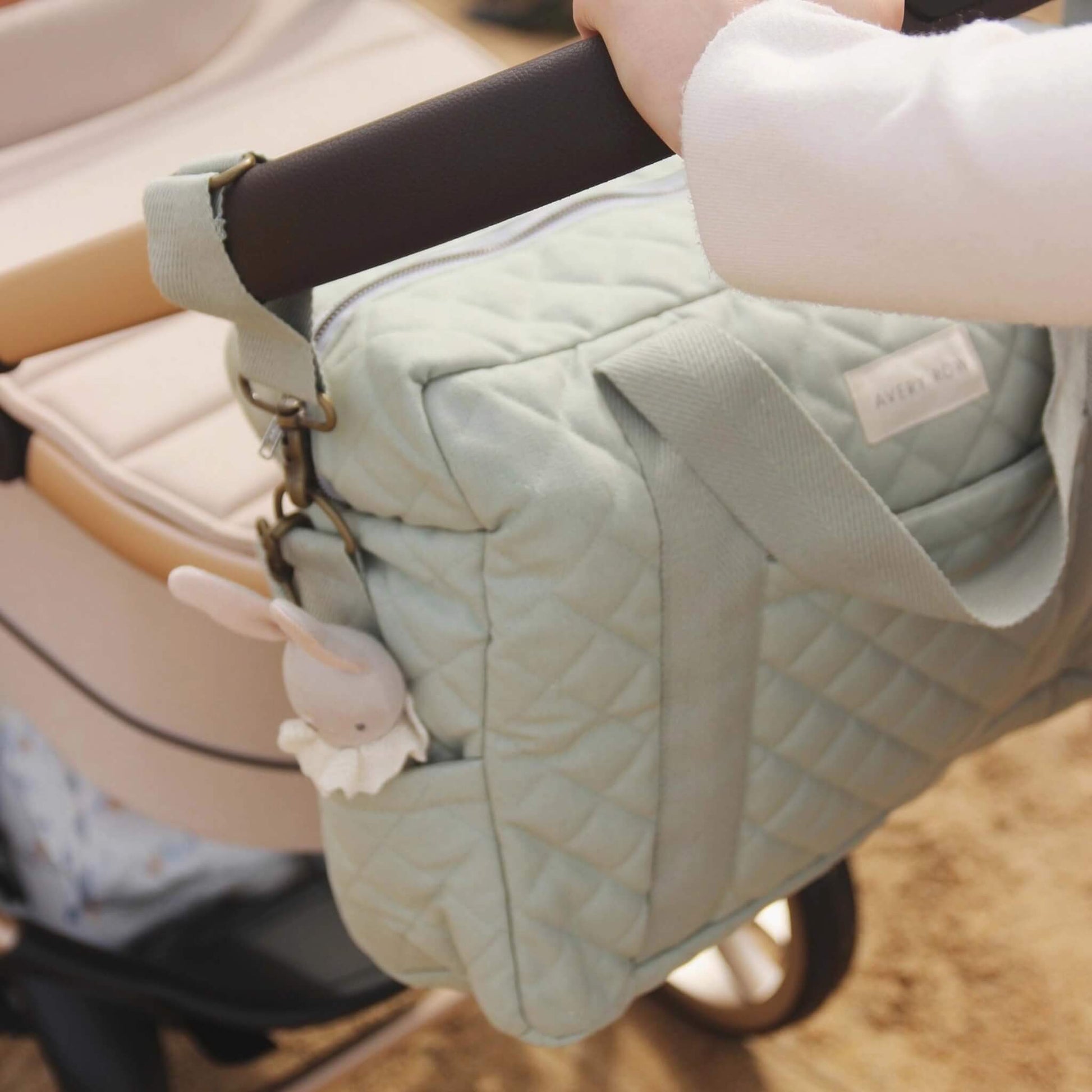 Sage green quilted changing bag clipped onto a pram handle using fabric straps and metal hooks, hanging neatly at the back of the stroller.