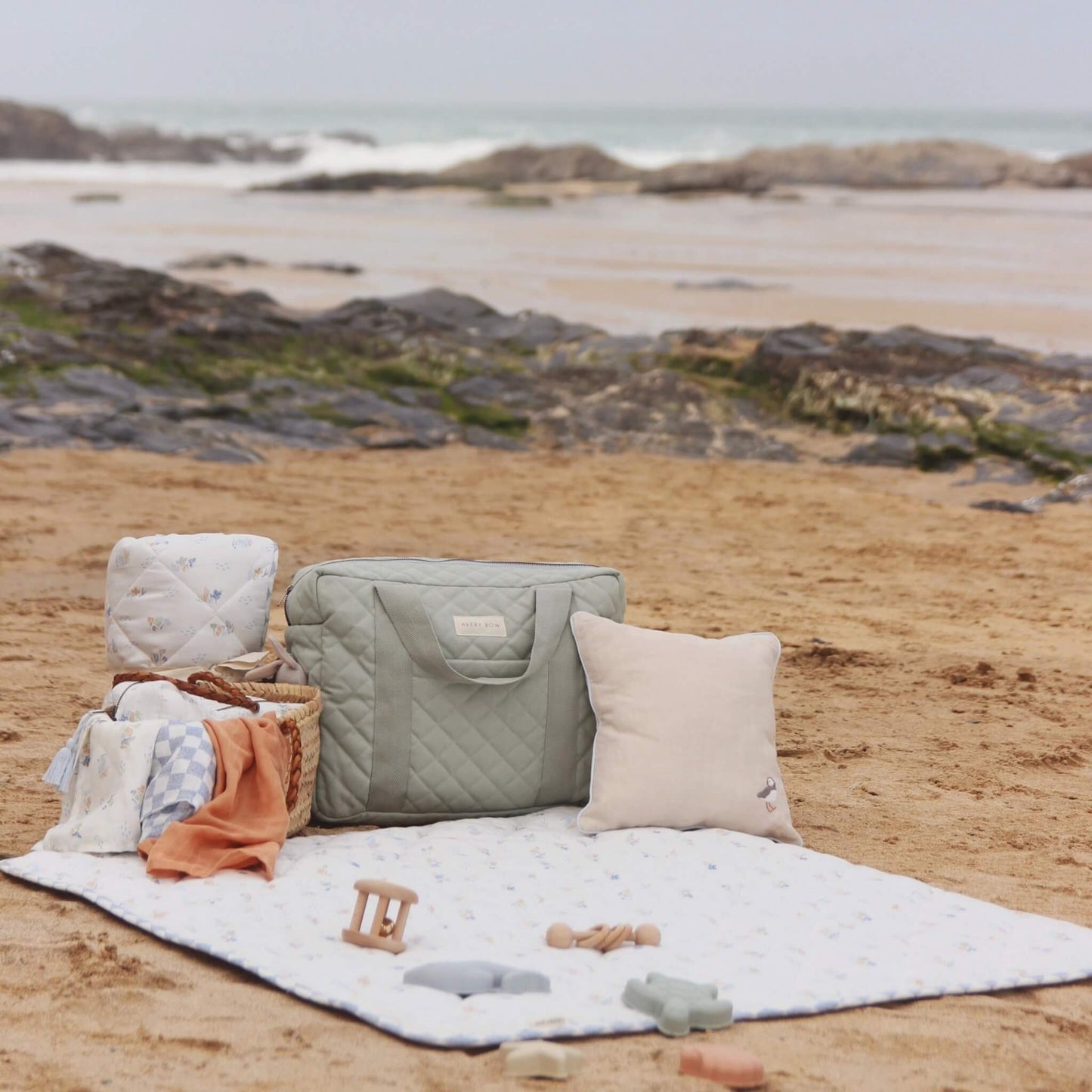 Quilted sage green nappy changing bag placed on a picnic blanket at a sandy beach with baby essentials, cushion and toys arranged around it.