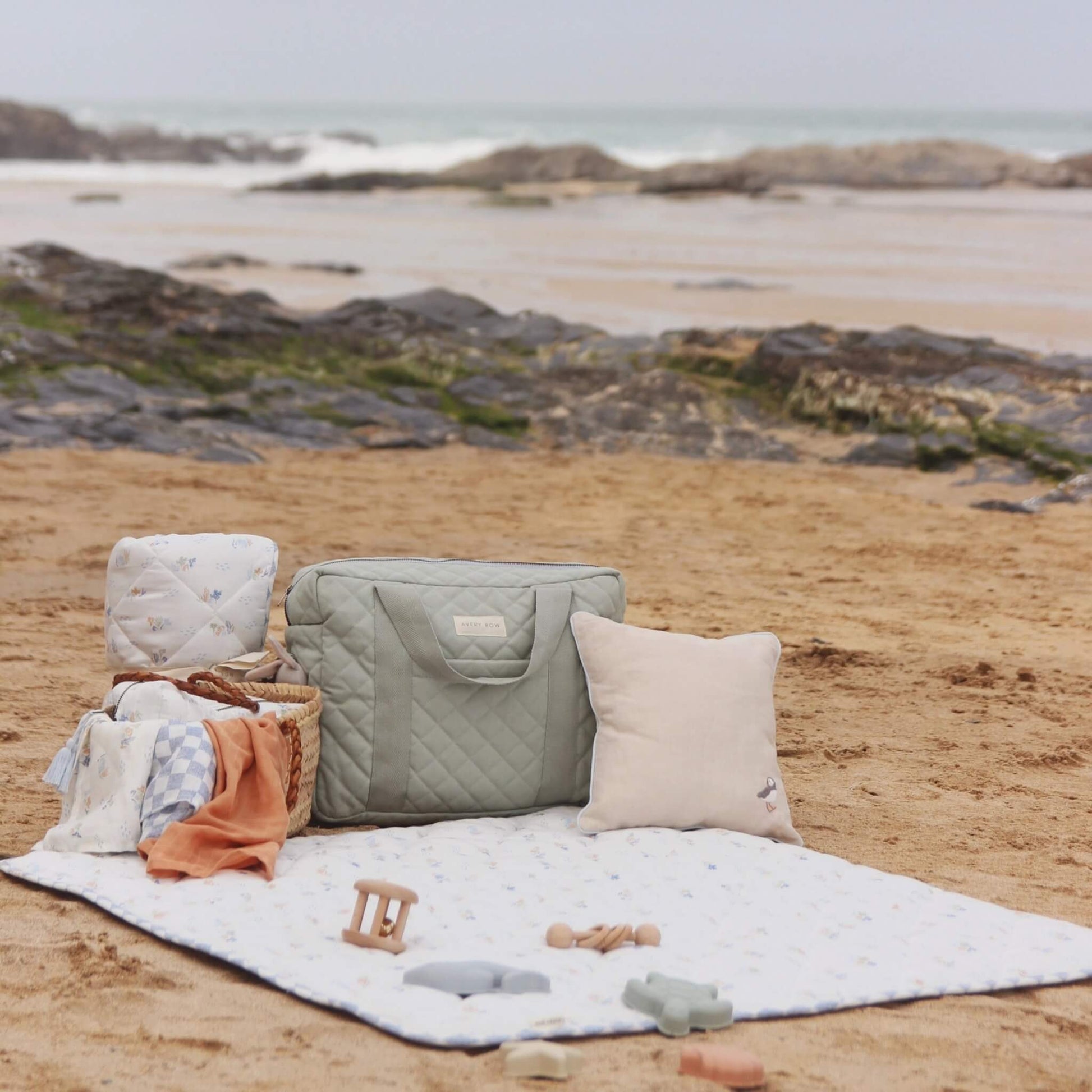 Quilted sage green nappy changing bag placed on a picnic blanket at a sandy beach with baby essentials, cushion and toys arranged around it.