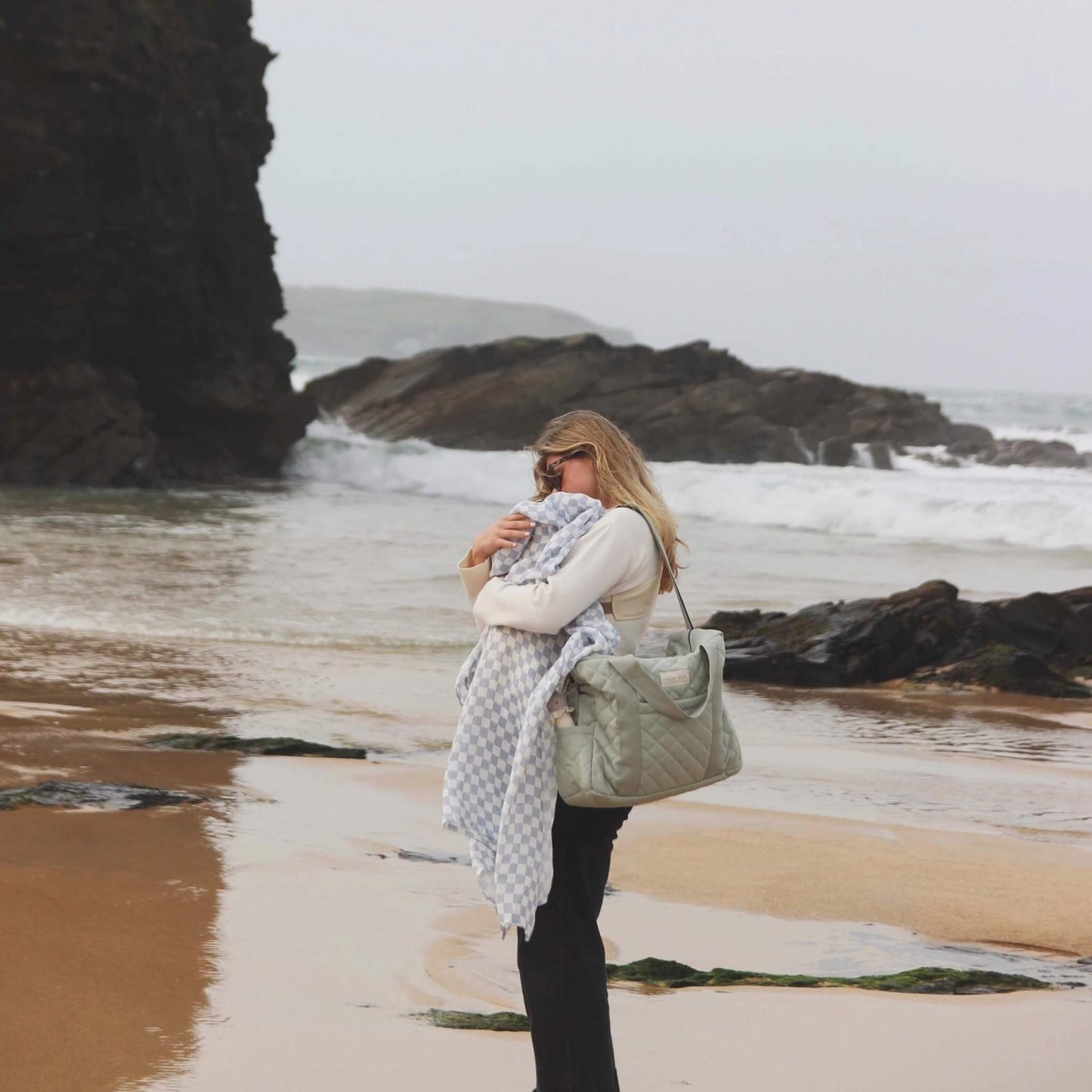 Woman carrying a soft quilted sage green changing bag over her shoulder at the beach while holding a blanket, showing everyday carry style.