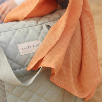 Close-up of quilted sage green cotton changing bag with stitched diamond pattern and woven label, partially covered by a soft rust coloured muslin cloth.