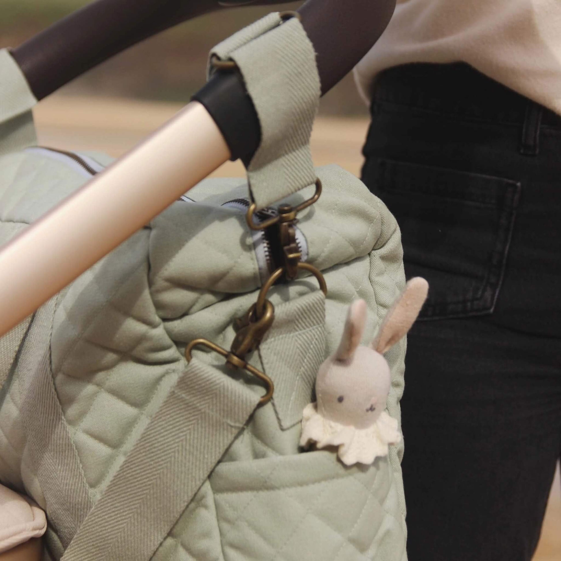 Quilted sage green changing bag hanging from a stroller handle using fabric straps and brass clips, with a small soft bunny toy tucked into a side pocket.