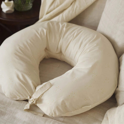 A cream nursing pillow resting on an armchair, showing its padded curved shape and soft cotton cover.